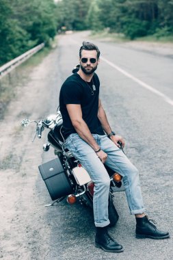 handsome motorcyclist in sunglasses sitting on black motorcycle on road near green forest clipart