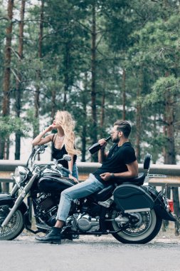 yeşil orman yakınındaki yolda şişeden alkol içen adam siyah motosiklet üzerinde oturan bisikletçilerin genç çift yan görünümü