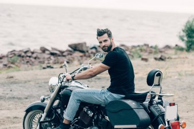 handsome bearded motorcyclist on black motorcycle at sandy beach near sea clipart