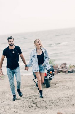 kumlu plajda siyah motosiklet yakınında yürüyen ve el ele tutuşan bisikletçilerin genç çift