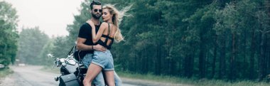 young sexy couple of motorcyclists embracing near black motorcycle on road near green forest, panoramic shot clipart