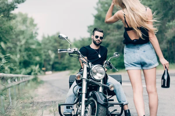 黒いオートバイのサングラスでハンサムなボーイフレンドの近くにアルコールのボトルを持って道路を歩いている若い女性のバックビュー — ストック写真