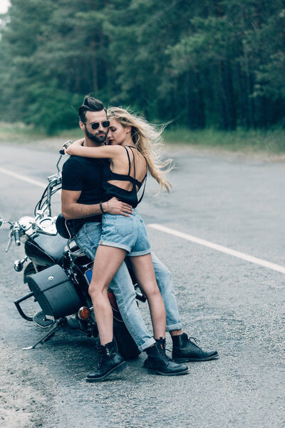 молодая сексуальная парочка мотоциклистов, обнимающихся возле черного мотоцикла на дороге возле зеленого леса
