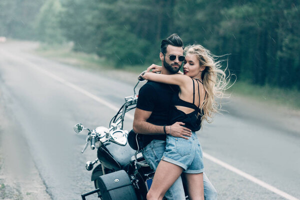 молодая сексуальная парочка мотоциклистов, обнимающихся возле черного мотоцикла на дороге возле зеленого леса
