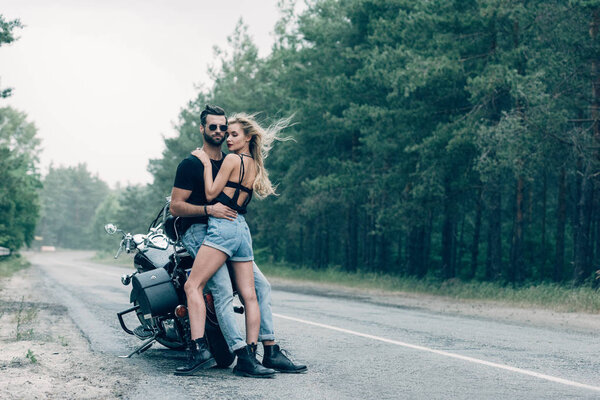 молодая сексуальная парочка мотоциклистов, обнимающихся возле черного мотоцикла на дороге возле леса
