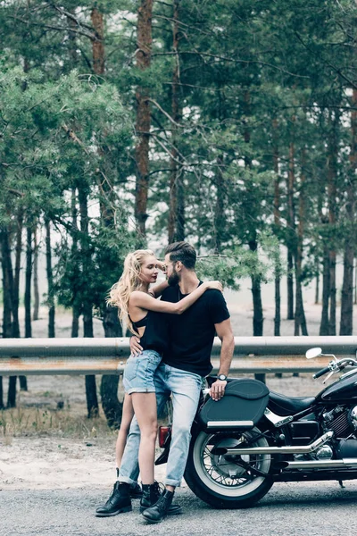 黒いオートバイと緑の森の近くで抱きしめるバイカーのセクシーな若いカップル — ストック写真