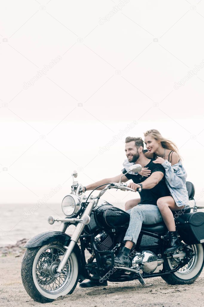 happy young couple of bikers riding black motorcycle at sandy beach