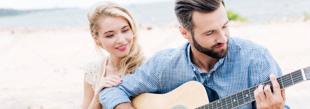 smiling beautiful young woman sitting near boyfriend with acoustic guitar at beach near sea, panoramic shot