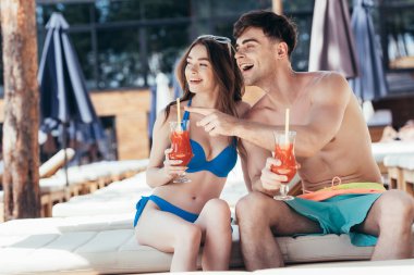 cheerful young man pointing with finger while sitting on deck chair near attractive girlfriend  clipart