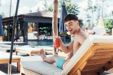 cheerful young man smiling at camera while sitting on deck chair with glass of refreshing beverage clipart