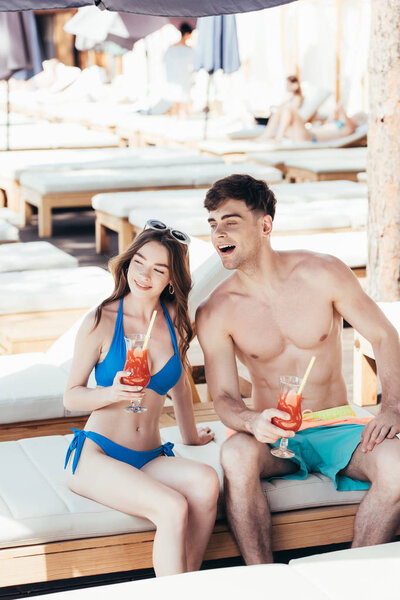 cheerful young couple sitting on deck chair with glasses of refreshing beverage and smiling while looking away