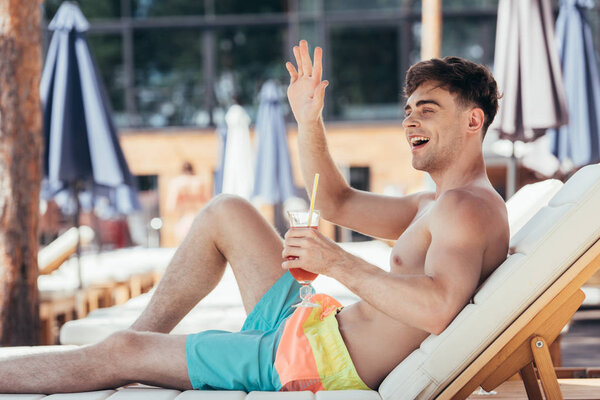 cheerful young man waving looking away and waving hand while relaxing on chaise lounge with glass of refreshing drink