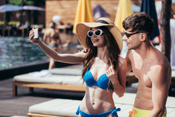 cheerful young woman in sunglasses and straw hat taking selfie with handsome boyfriend