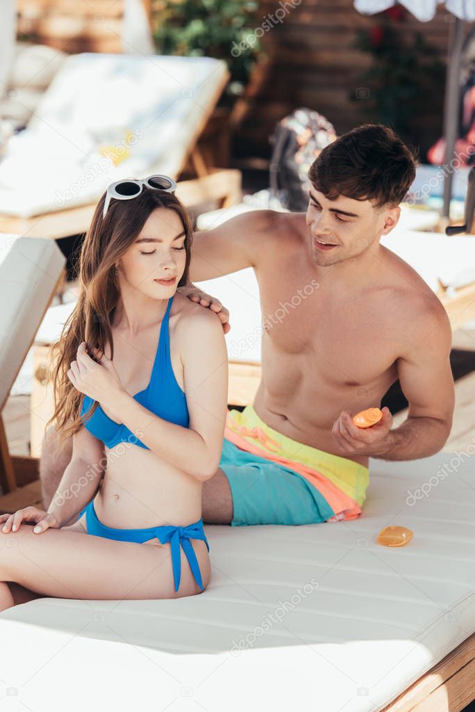 attentive young man applying sunscreen cream on shoulder of smiling girlfriend