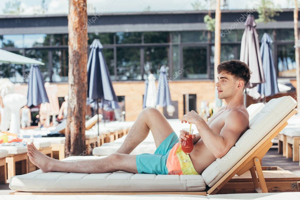 handsome young man looking away while relaxing on deck chair with glass of refreshing drink