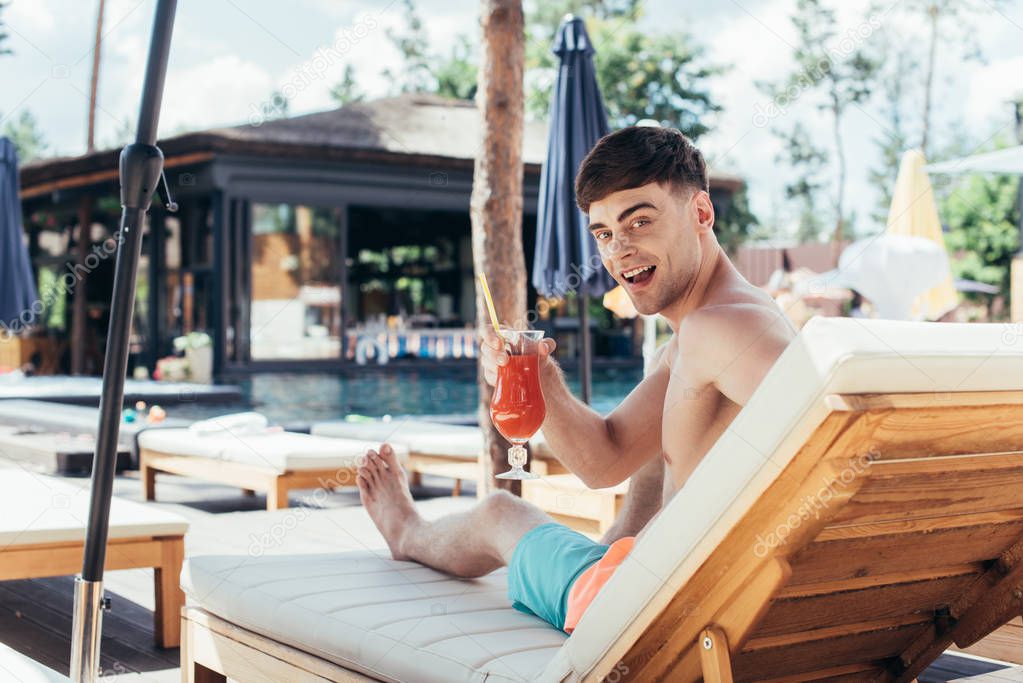 cheerful young man smiling at camera while sitting on deck chair with glass of refreshing beverage