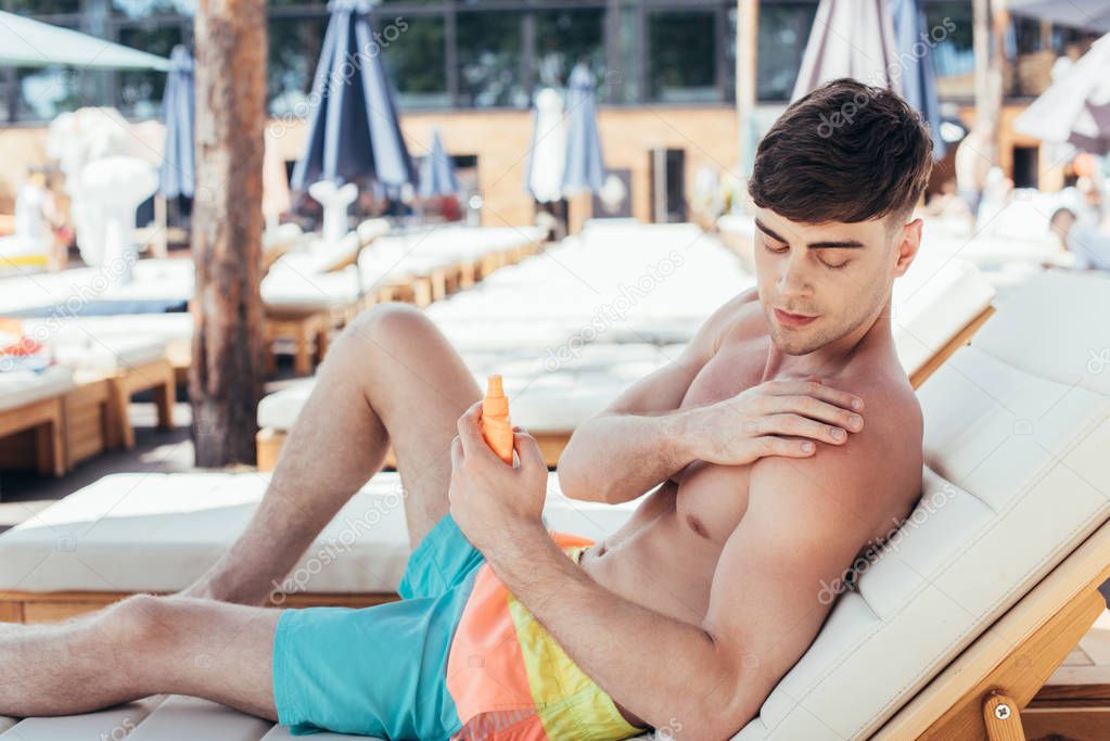 handsome young man applying sunscreen lotion while sitting on chaise lounge