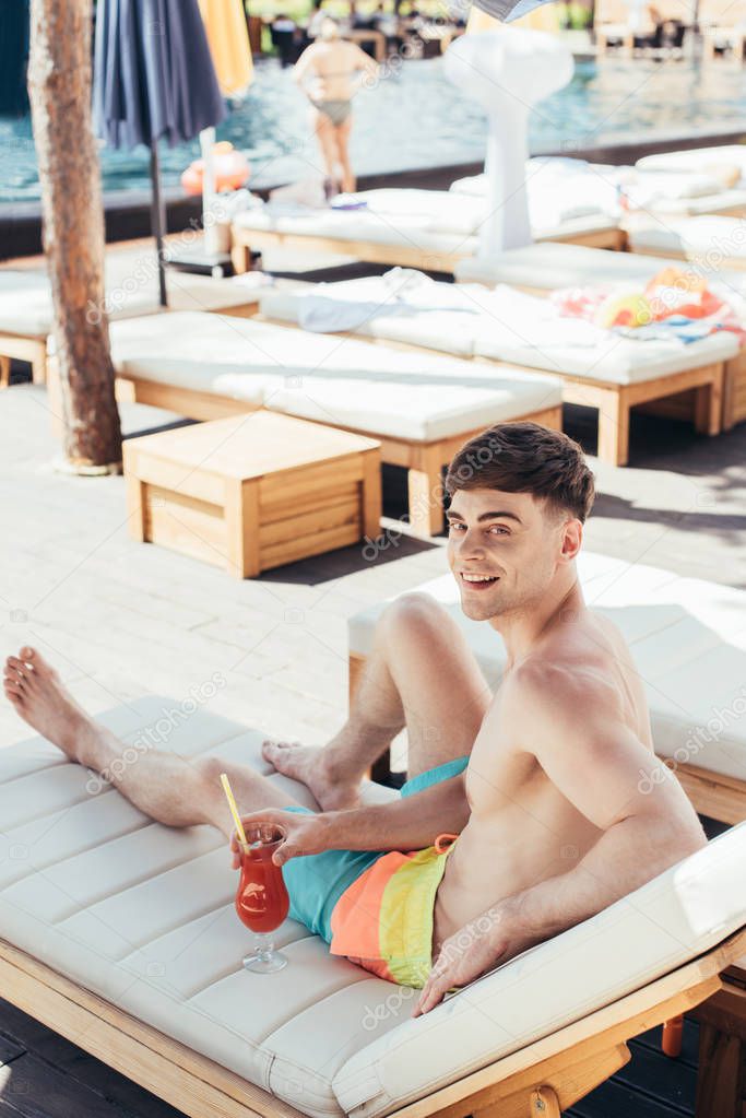handsome young man smiling at camera while resting on deck chair with glass of refreshing beverage