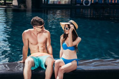 yüzme havuzu yakınında otururken birbirlerine bakarak gülümseyen genç çift