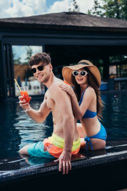 mutlu genç çift gülümseyerek ve havuz başında otururken kameraya bakarak