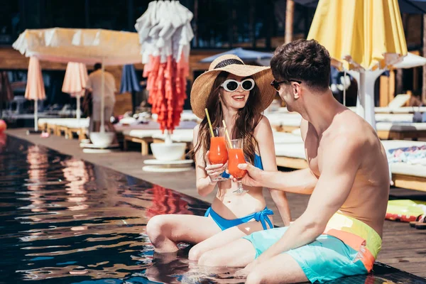 陽気な若いカップルは プールサイドに座っている間 さわやかな飲み物で眼鏡を鳴らします — ストック写真