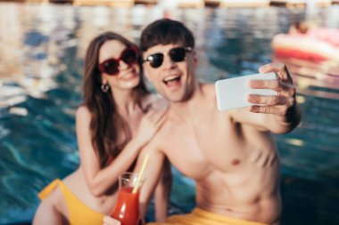 havuz başında otururken selfie çeken neşeli genç çiftin seçici odağı