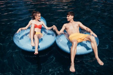 yüzme havuzunda yüzme halkaları üzerinde dinlenirken el ele tutuşan mutlu genç çift