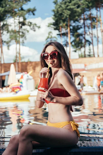 水着とサングラスを着た可愛い若い女性が爽やかな飲み物を飲み プールサイドに座りながらカメラを見ている — ストック写真