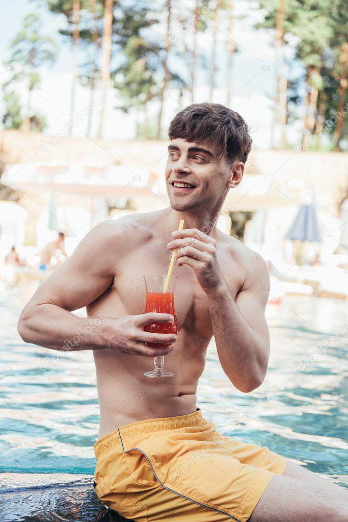 cheerful young man looking away while sitting at poolside with glass of refreshing drink