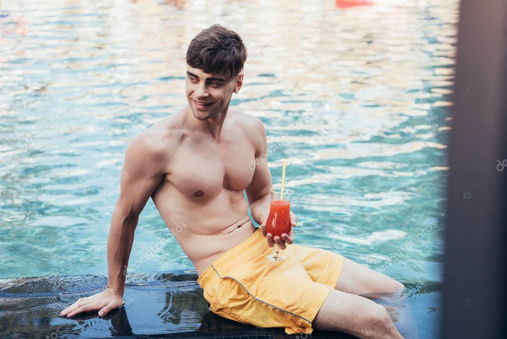 selective focus of cheerful young man looking away while relaxing at poolside with glass of refreshing drink