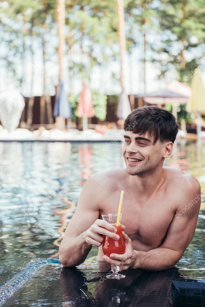 handsome young man looking away while relaxing in swimming pool with glass of refreshing drink
