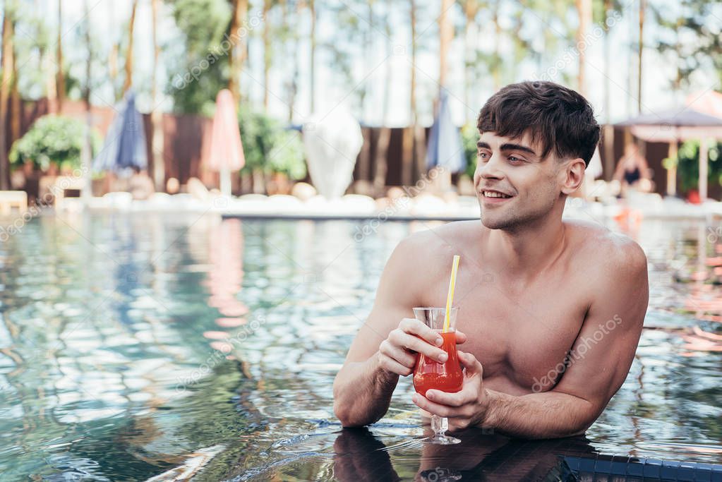 smiling young man looking away while relaxing in swimming pool with glass of refreshing drink