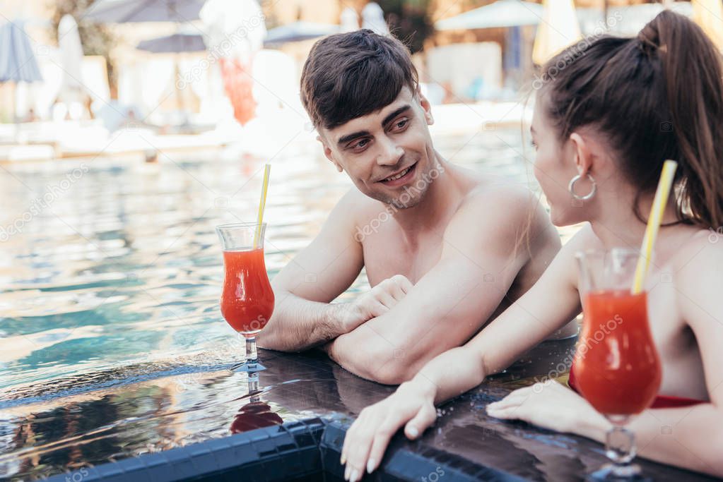 cheerful young couple looking at each other while resting at poolside with glasses of refreshing drink