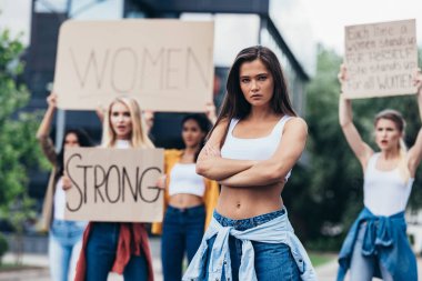 sokakta feminist sloganlar taşıyan pankartlar tutan kadınların yanında kolları kapalı duran ciddi feminist ayakta