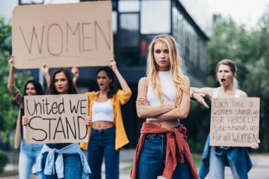 serious woman standing with arms crossed near women holding placards with feminist slogans on street clipart