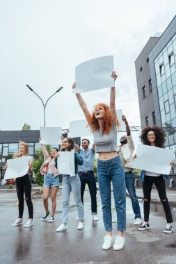 çok kültürlü insanların yakınında boş tahta tutan mutlu kızıl kız seçici odak 