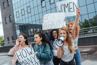 selective focus of emotional girl holding megaphone and gesturing near multicultural women holding placards on meeting  clipart