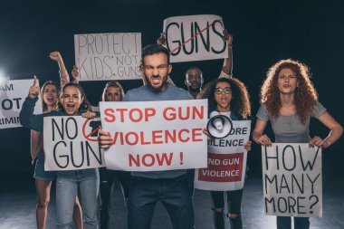 angry man holding placard with stop gun violence now lettering near multicultural people on black  clipart