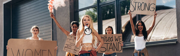 panoramic shot of five screaming multiethnic feminists holding smoke bomb, loudspeaker and placards with feminist slogans