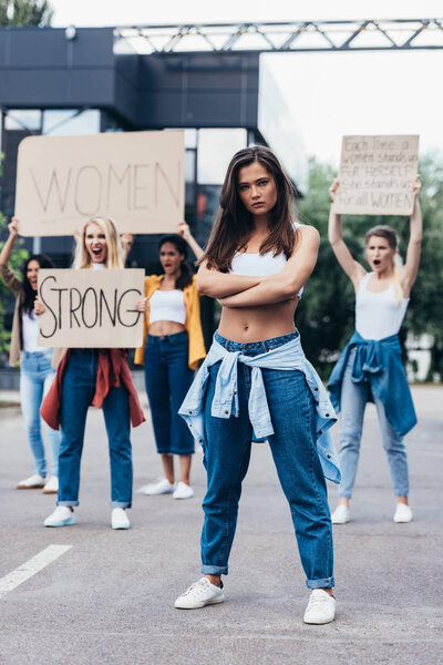 full length view of serious feminist standing with arms closed near women holding placards with feminist slogans on street