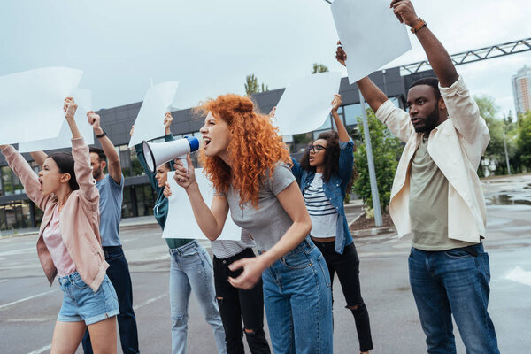 emotional redhead girl screaming in megaphone near multicultural people 