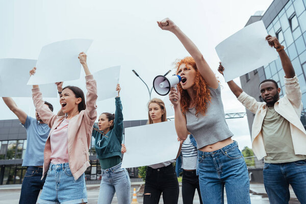 emotional redhead woman screaming in megaphone near multicultural people 