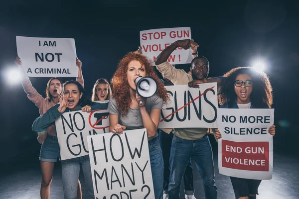 redhead girl holding placard with how many more lettering and screaming in megaphone near multicultural people on black