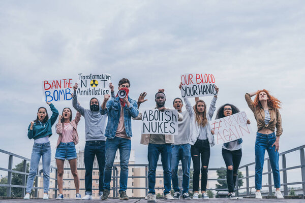 Человек с шарфом на лице держит мегафон рядом с мультикультурными людьми с плакатами
 