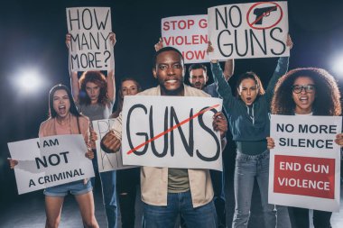 emotional african american man holding placard with guns lettering near group of people on black  clipart