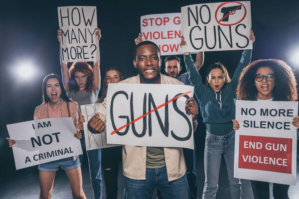 emotional african american man holding placard with guns lettering near group of people on black 