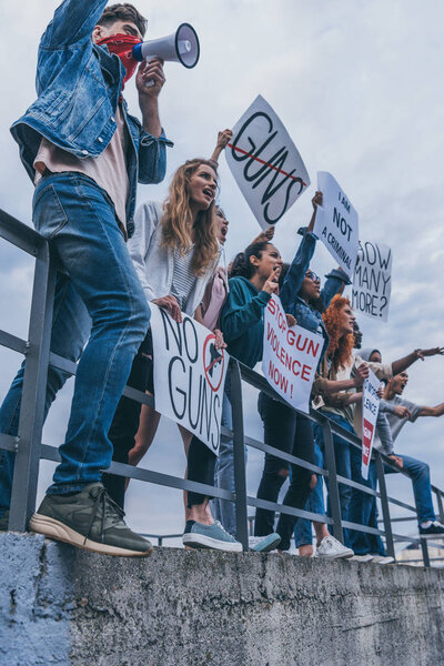 low angle view of man with scarf on face screaming in megaphone near multicultural people with lettering on placards 