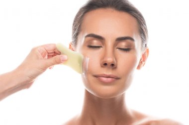 partial view of cosmetologist applying face cream and using gua sha scraper isolated on white clipart