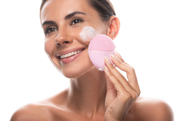 nude young woman with cream on face using facial cleansing brush isolated on white