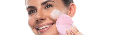 panoramic shot of smiling woman with cream on face using facial cleansing brush isolated on white clipart